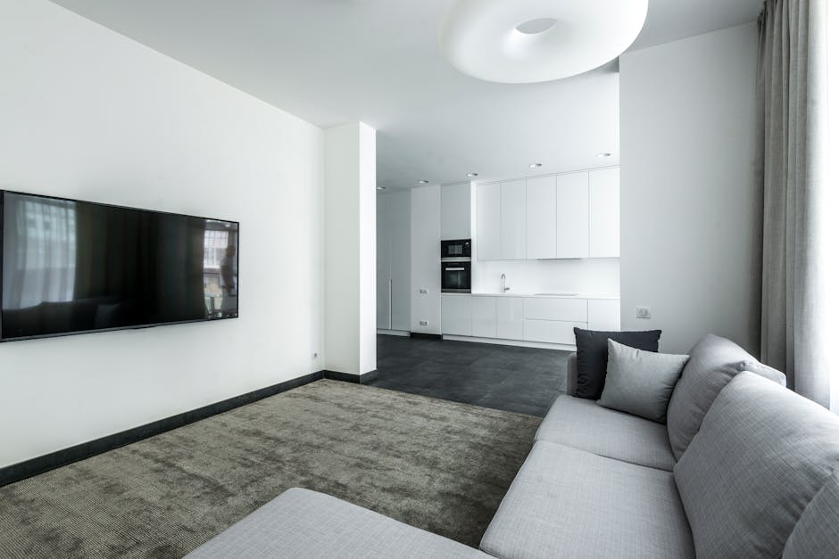 A modern, minimalist living room with a light grey fabric sofa accented with various dark and light grey throw pillows, positioned along the left wall. The room features a beige textured carpet coveri