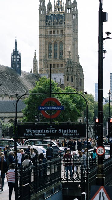 A view of Westminster Station in London featuring the red underground sign, with a group of pedestrians walking on the sidewalk near a black metal fence. In the background, the London Eye Ferris wheel